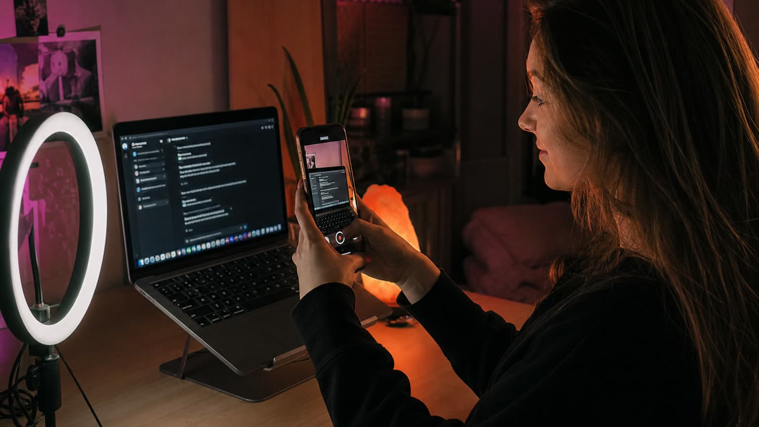 A creator with long hair holds her phone vertically to film her own laptop screen in a dim bedroom studio; the laptop shows a dark chat interface, a ring light glows on the left and a salt lamp burns orange behind.